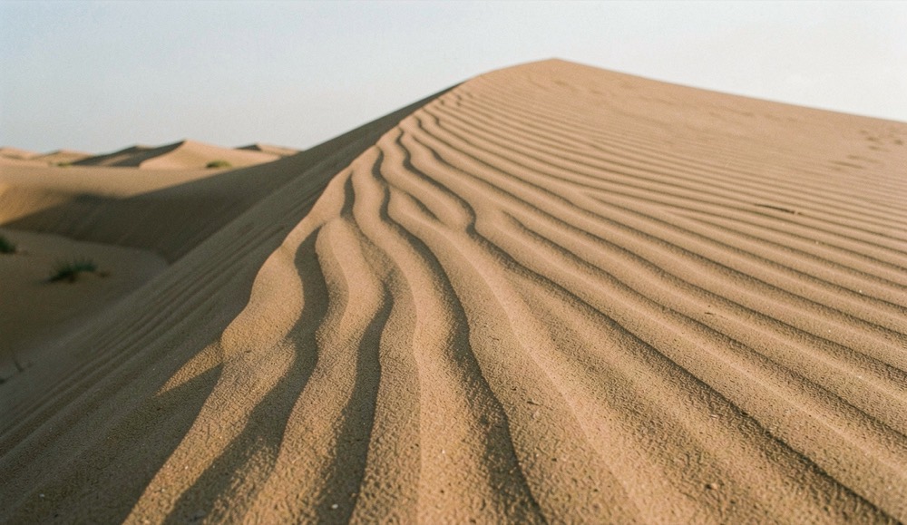 Crête rocheuse au coucher du soleil, chaque bloc projette une ombre longue vers la gauche du cadre, la poussière de la piste reste en suspension dans l'air chaud