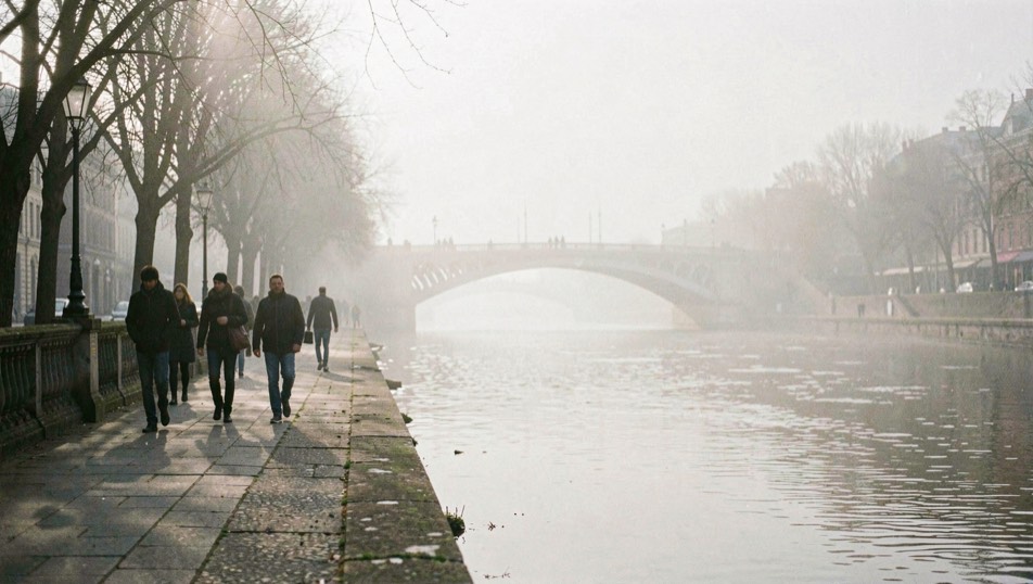 Quai le long d'un fleuve dans une brume légère, silhouettes de passants et arche de pont qui pâlit au fond