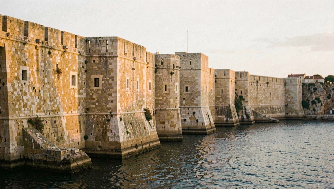 Bastions de pierre dorée surplombant un port naturel, la lumière rasante du soir accentue les assises taillées et les meurtrières