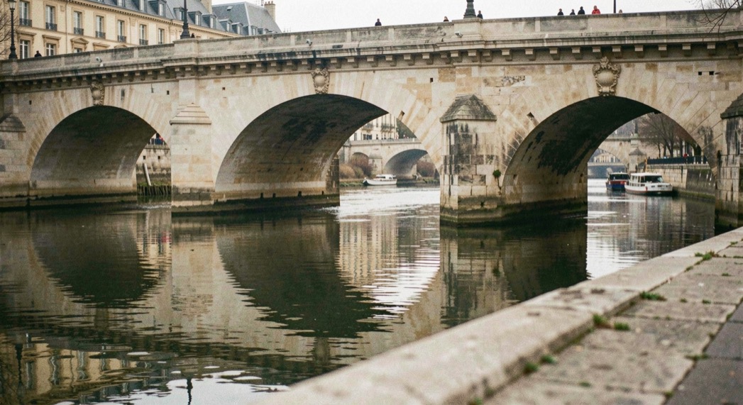 Pont de pierre à arches se reflétant dans la Seine à l'heure bleue, les lampadaires allumés dessinent une ligne de points dorés sur un ciel encore bleu